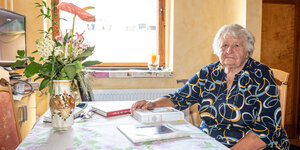 Anastasia Gulaj sitzt an einem Tisch vor dem Fenster, ein Buch liegt vor ihr und ein Strauß Blumen steht in einer Vase