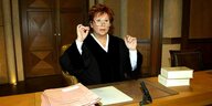 Barbara Salesch sitzt in Richterinnenkleidung (schwarzer Kittel) an einem Holztisch im Gerichtssaal. Sie ist eine Frau mittleren alters mit kurzen roten Haaren und Brille. Sie schaut schockiert und hebt beide Hände auf Kopfhöhe. Auf dem Tisch: Ordner und