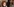 Eine Frau schminkt sich am Spiegel, eine andere mit Brille und lockigem kurzen Haar schaut eine Person hinter der Kamera an
