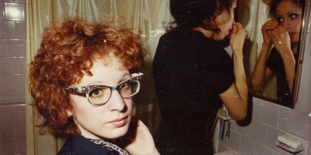 Eine Frau schminkt sich am Spiegel, eine andere mit Brille und lockigem kurzen Haar schaut eine Person hinter der Kamera an
