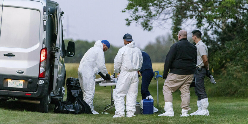 Menschen teilweise in weißen Schutzanzügen stehen auf einer Wiese zusammen um einen Tisch herum