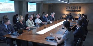 Etwa ein Dutzend Menschen sitzen an einem langen hölzernen Konferenztisch. Manche der Personen haben Blöcke oder Bildschirme vor sich. Hinter den Menschen noch eine Reihe Menschen auf Stühlen.