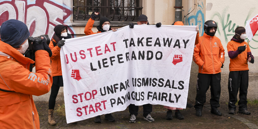 Lieferando-Fahrer:innen demonstrieren mit einem Banner für bessere Löhne und gegen Kündigungen.