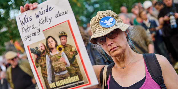 Eine Frau mit Anglerhut hält ein Spiegel-Cover mit dem Titel "Die Olivgrünen" hoch, darüber der Titel "Was würde Petra Kelly dazu sagen?"