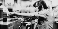Schwarz-weißes Video Still aus dem Film "Sing, Iris, Sing": Eine mit einem Hemd bekleidetet Frau steht in einer Fabrik an einer Kurbelbank und arbeitet