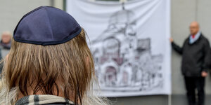 Ein Mann mit einer Kippa nimmt in Hamburg an einer Mahnwache Teil. Im Hintergrund wird eine Zeichnung der Bornplatz-Synagoge gezeigt.