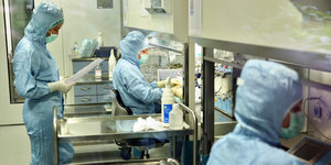 Mitarbeiterinnen mit Mundschutz in hellblauen Overals stellen im Labor Medikamente her.