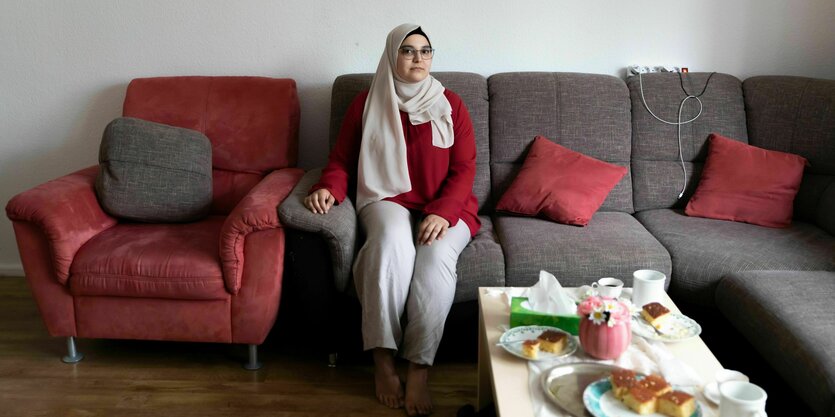 Bayan Alkhatib sitzt auf einem grauen Sofa, sie trägt einen roten Pullover und einen hellen Hijab