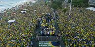 An einem Strand steht eine in blau-gelb gekleidete Menschenmenge. In der Mitte spricht Jair Bolsonaro in ein Mikrofon