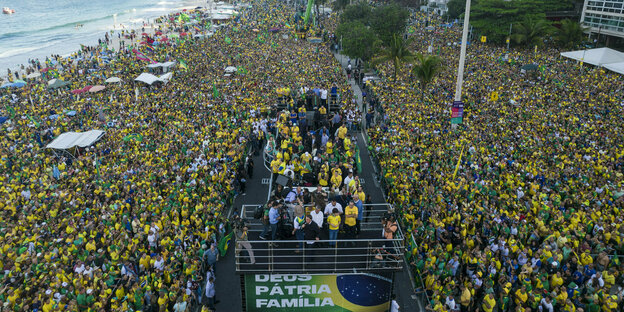 A crowd dressed in blue and yellow is standing on a beach.  In the middle, Jair Bolsonaro speaks into a microphone
