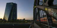Die EZB leuchtet im letzten Licht des Tages während auf der benachbarten Eisenbahnbrücke ein vorbeifahrender Zug Lichtstreifen durch die Dunkelheit zieht