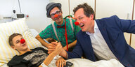 Zwei Männer sitzen an einem Krankenhausbett und lachen gemeinsam mit einem kranken Jungen, alle haben rote Clownsnasen auf
