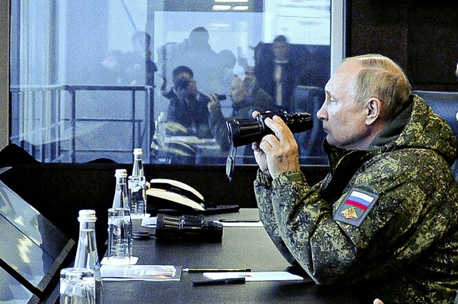 Wladimir Putin sitzt in Uniform an einem Tisch und hält ein Fernglas in den Händen