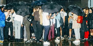 Es regnet, Menschen stehen dircht gedrängt mit Regenschirmen unter einem Vordach