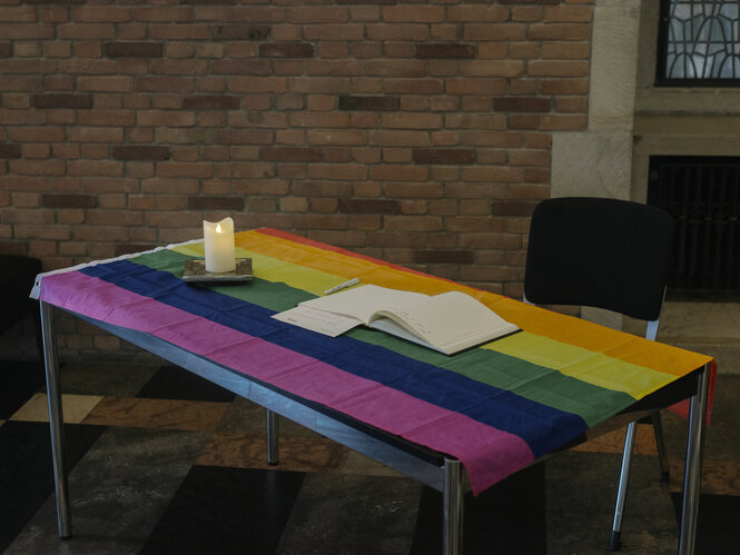 Auf einem Tisch liegt eine Decke in Regenbogenfarben, darauf steht eine Kerze und ein Kondolzenbuch