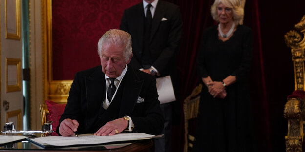 King Charles III  signs an oath in the presence of his son, William, Prince of Wales, and his wife, Queen Camilla
