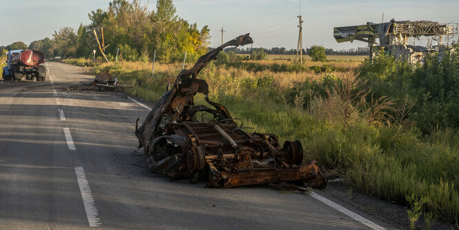 Zerstörte Fahrzeuge stehen auf einer Landstraße, im Hintergrund ein schwer beschädigtes Gebäude