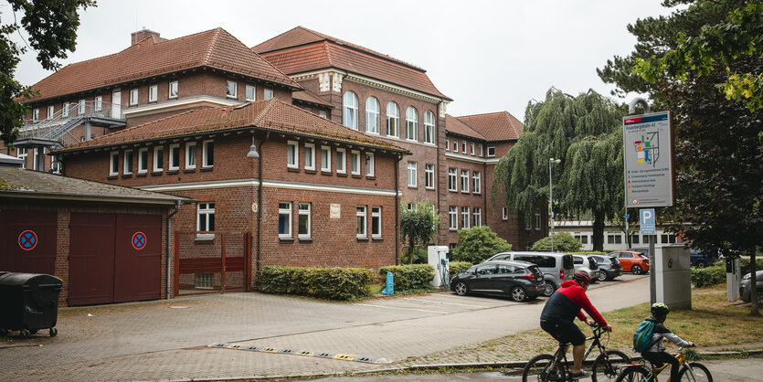 Zwei Menschen fahren mit einem Fahrrad an einem Parkplatz vor einem großen Gebäude in roten Klindersteinen vorbei