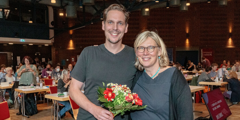 Ein Mann und eine Frau haltenlächelnd gemeinsam einen Blumenstrauß in der Hand, im Hintergrund sitzen Delegierte an Tischen