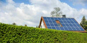 Ein Einfamilienhaus mit Solarzellen auf dem Hausdach steht hinter einer grünen Hecke.