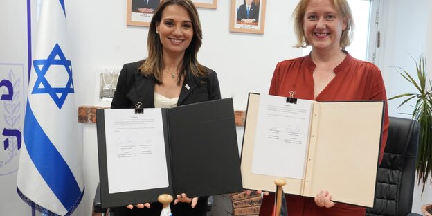Two women hold documents for the camera, the Israeli flag on the left
