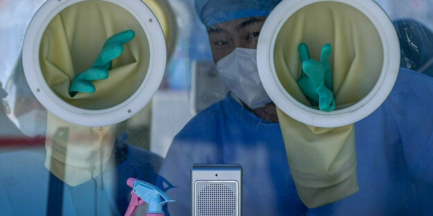A medical worker in protective gear looks through a window behind a handle slot