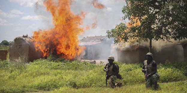 Zwei Soldaten auf einer Wiese vor einem brennenden Haus