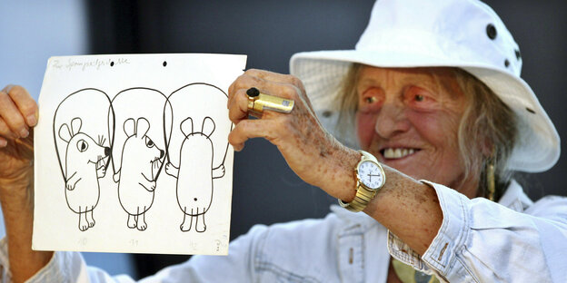 An old woman with a hat and gold jewelry shows drawings of the mouse