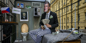 Ein Mann mit Andrej Babiš Maske sitzt in einer Gefängniszelle, Tresor, Geld und Clo hinter Gittern