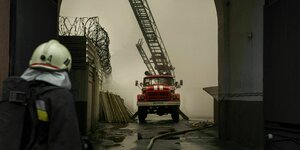 Ein Feuerwehrauto mit ausgefahrener Leiter steht in einem verrauchten Durchgang, im Vordergrund steht ein Feuerwehrmann in Montur