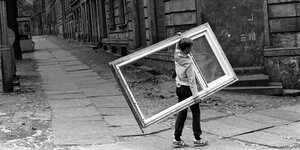 Ein junger Mann trägt einen großen Fensterrahmen durch das graue Ostberlin der Vorwendezeit