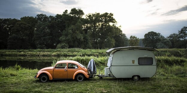 Ein orangfarbener VW Käfer und ein Wohnwagen in einer Auenlandschaft
