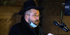 Ein Rabbiner mit schwarzem Hut und runtergezogenen Mundschutz spricht ins Mikrofon
