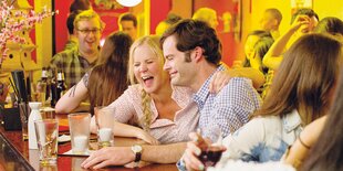 HauptdarstellerInnen Amy Schumer und Bill Hader sitzen an einer Bar und lachen, sie hat einen Arm um seine Schulter gelegt