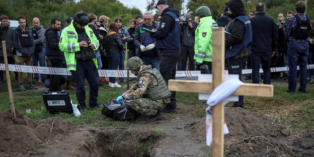 Soldaten und Menschen und zwei Leute in Warnweste vor einer ausgehobenen Grube, daneben ein Kreuz aus Holzlatten