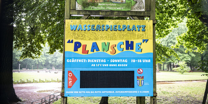 Ein Schild mit der Aufschrift "Wasserspielplatz Plansche" weißt auf die Öffnungszeiten des Spielplatzes hin