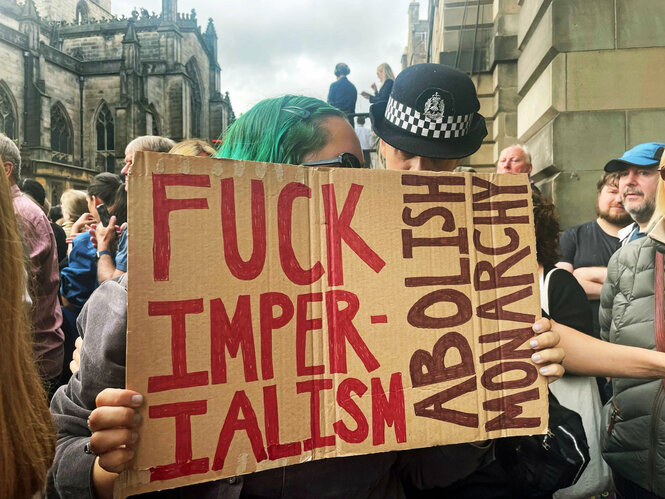 Eine Polizistin spricht mit einer jungen Frau, die ein Schild mit der Aufschrift "Fuck Imperialism - Abolish monarchy" in der Hand hält