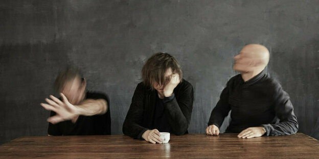 Das Michael Wollny Trio sitzt vor einer dunkelgrauen Wand an einem Holztisch, die Gesichter der drei Mitglieder sind verwischt