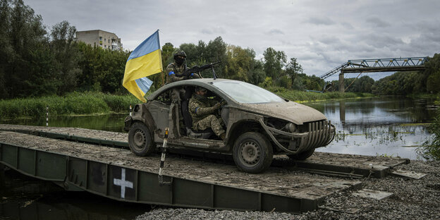 Ukrainian soldiers sit in a car with a Ukraine flag and cross a river.