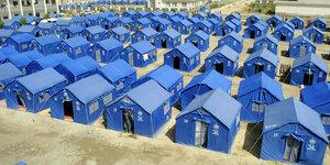 Blaue Zelte, die wie Häuschen aussehen stehen aufgereiht auf einem staubigen Platz umgeben von Neubauten