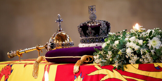 On a coffin covered with flags and flowers lie a scepter, an orb and a crown on a pillow