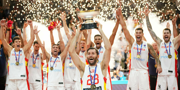 Eine medaillenbehangene Basketballmannschaft in weißen Leibchen jubelt, der Kapitän hält einen Pokal hoch, im Hintergrund Feuerwerk