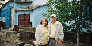Ein älteres Ehepaar steht Arm in Arm vor einem blau gestrichenen dörflichen Haus