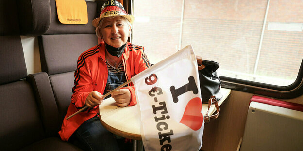 Eine Frau fährt Zug und hat eine Fahne in den Händen auf der geschrieben steht "I love 9-Euro-Ticket"