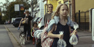 Szene aus „Petite nature“: Eine Familie läuft mit viel Gepäck und einem Hund eine Straße entlang, vorne rechts im Bild ist der zehnjährige Johnny zu sehen. Er trägt zwei mit Wasser gefüllte Plastikbeutel in der Hand, in denen Fische schwimmen, und eine große Kunststofftragetasche über der Schulter
