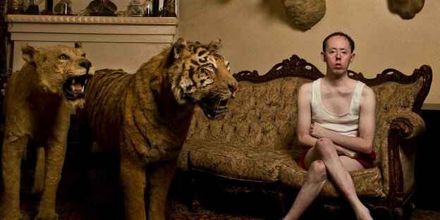 Actor Simon Laherty sits cross-legged in his underwear on a couch, with stuffed wild cats next to him