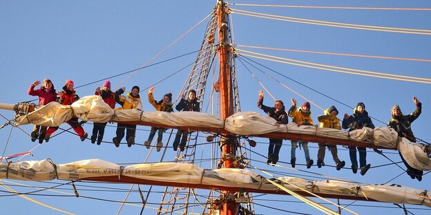 Jugendliche stehen auf dem Mastbaum eines Segelschiffs und winken