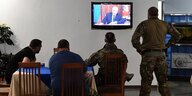 Vier Soldaten sitzen vor einem Fernsehschirm auf dem Putin zu sehen ist