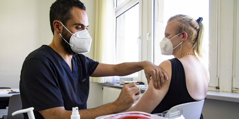 Zu sehen ist ein Arzt in blauem Arztkittel. Er impft gerade einer zweiten weiblich gelesenen Person in den Arm. Beide tragen eine Maske und sind nur mit ihrem Torso zu sehen.