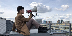 Ein Mann im Mantel sitzt mit einem Megafon am Mund auf einem Dach und schaut nach vorn.